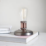 Endon Brackley industrial table lamp in aged pewter and copper with knurled E27 holder and exposed filament bulb, shown on books in a bright interior.