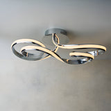 Endon Neve LED semi-flush ceiling light with swirling looped arms in polished chrome, mounted to a neutral ceiling above a light wall.