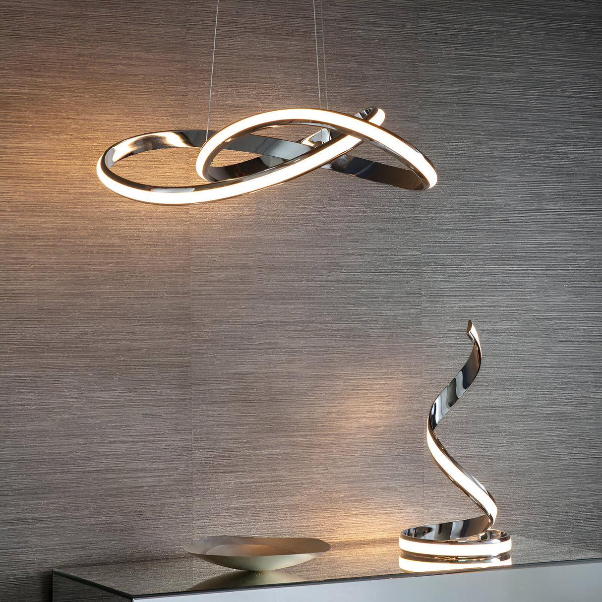 Endon Neve chrome LED pendant light with looping sculptural curves and white diffuser, shown lit above a matching spiral table lamp on a grey textured wall.