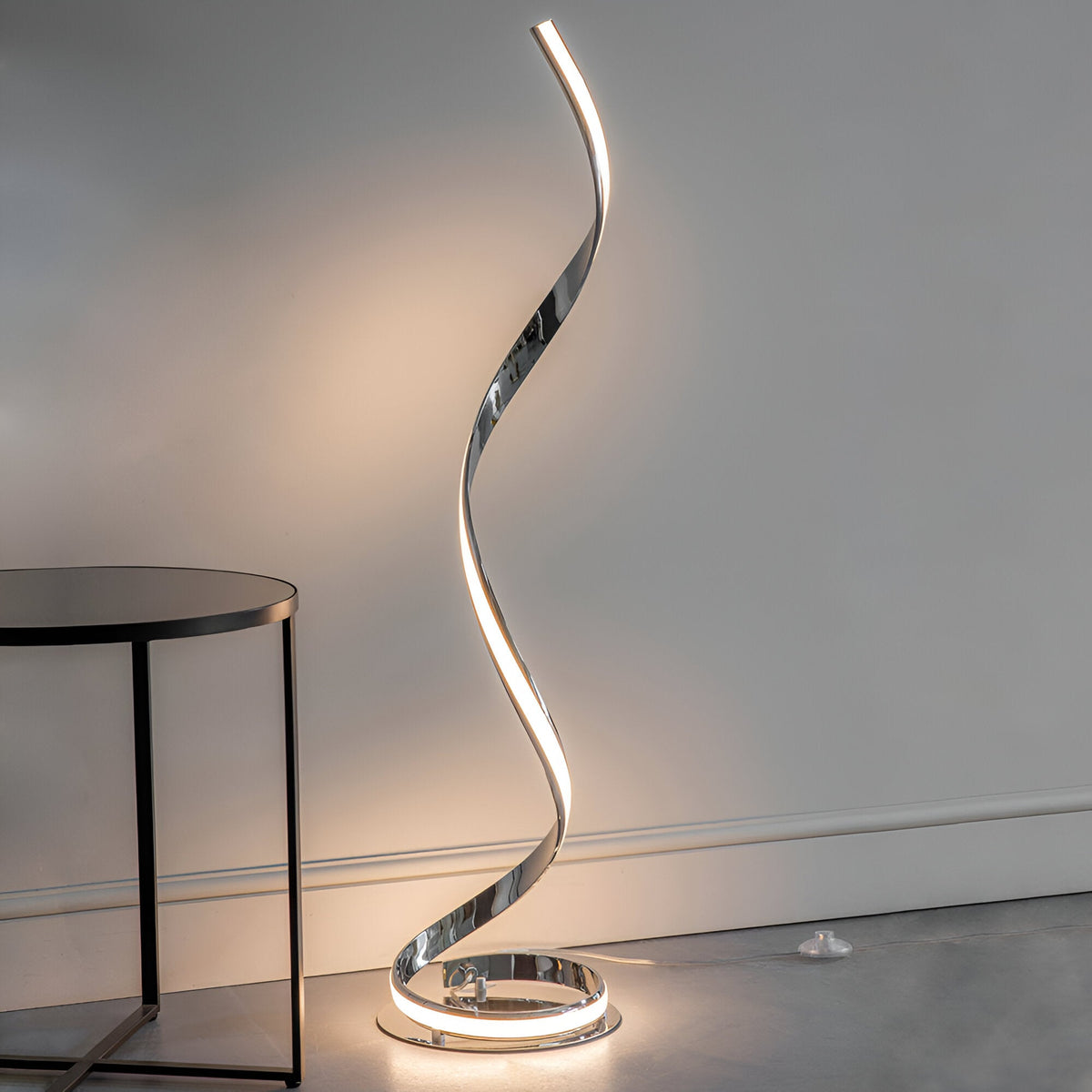 Neve Floor Lamp in polished chrome with an illuminated spiral curve and white diffuser, shown lit beside a black side table in a living room.