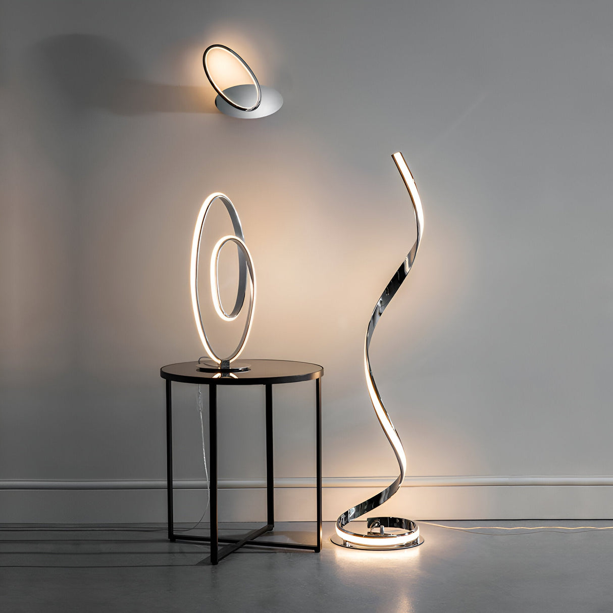 Neve Floor Lamp in polished chrome with an illuminated S-curve LED strip and white diffuser, shown lit beside a wall light and table lamp in a grey room.