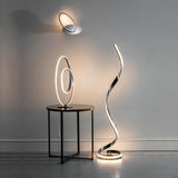 Neve Floor Lamp in polished chrome with an illuminated S-curve LED strip and white diffuser, shown lit beside a wall light and table lamp in a grey room.