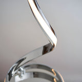 Close-up of the Neve Floor Lamp's spiralling chrome curve with a warm white LED diffuser, set against a plain light grey background.