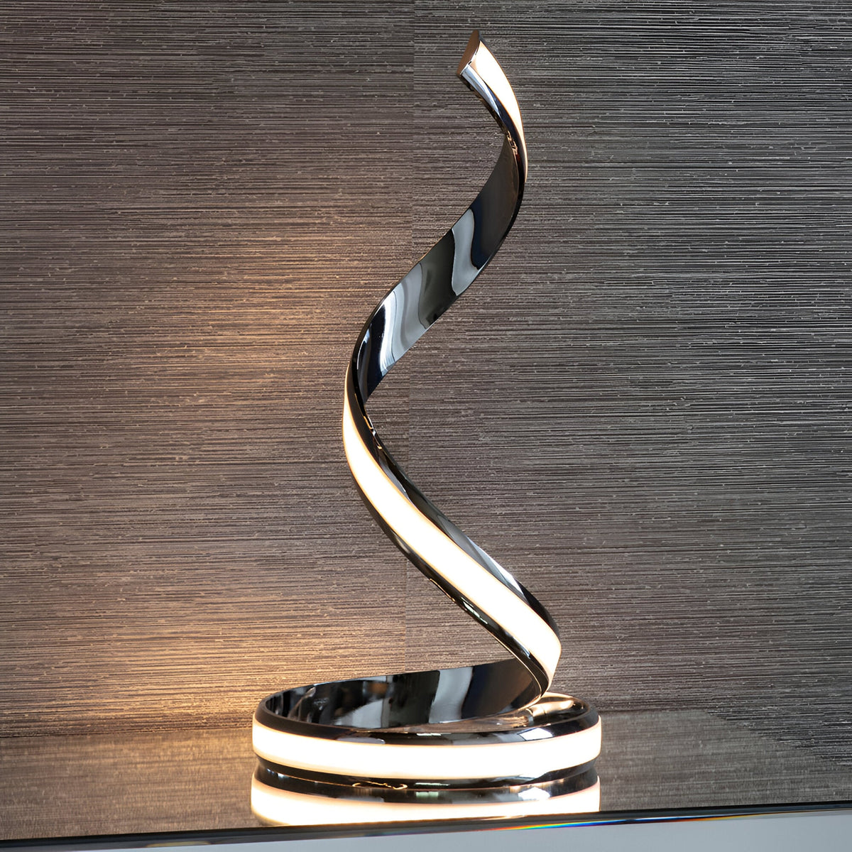 Endon Neve table lamp in polished chrome with a spiralling ribbon form and integrated LED diffuser, shown on a shelf against a textured grey wall.