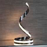 Endon Neve table lamp in polished chrome with a spiralling ribbon form and integrated LED diffuser, shown on a shelf against a textured grey wall.