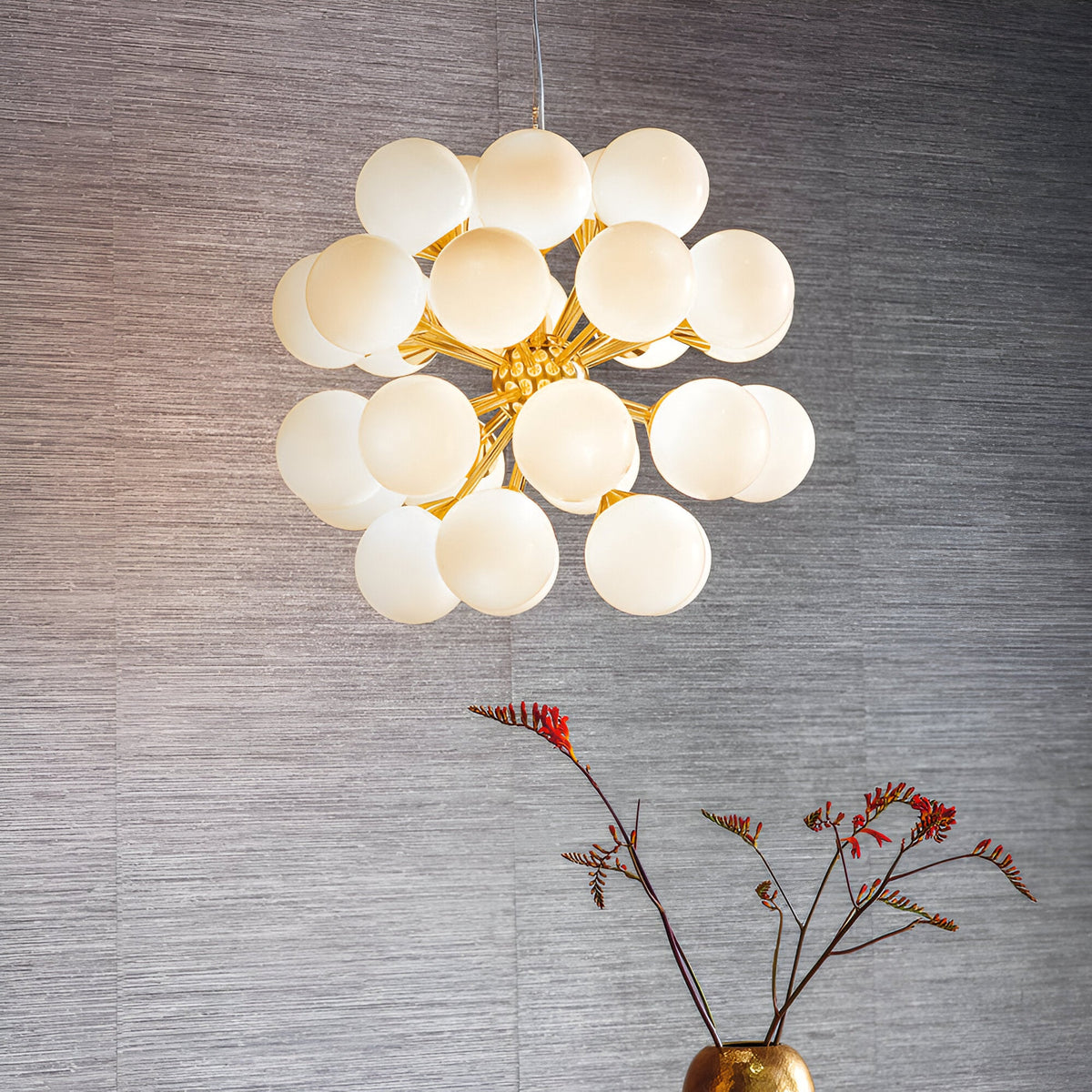 Fitzroy 28 Light Pendant in brushed brass with clustered gloss white opal glass globes, hung above a gold vase against a grey textured wall.