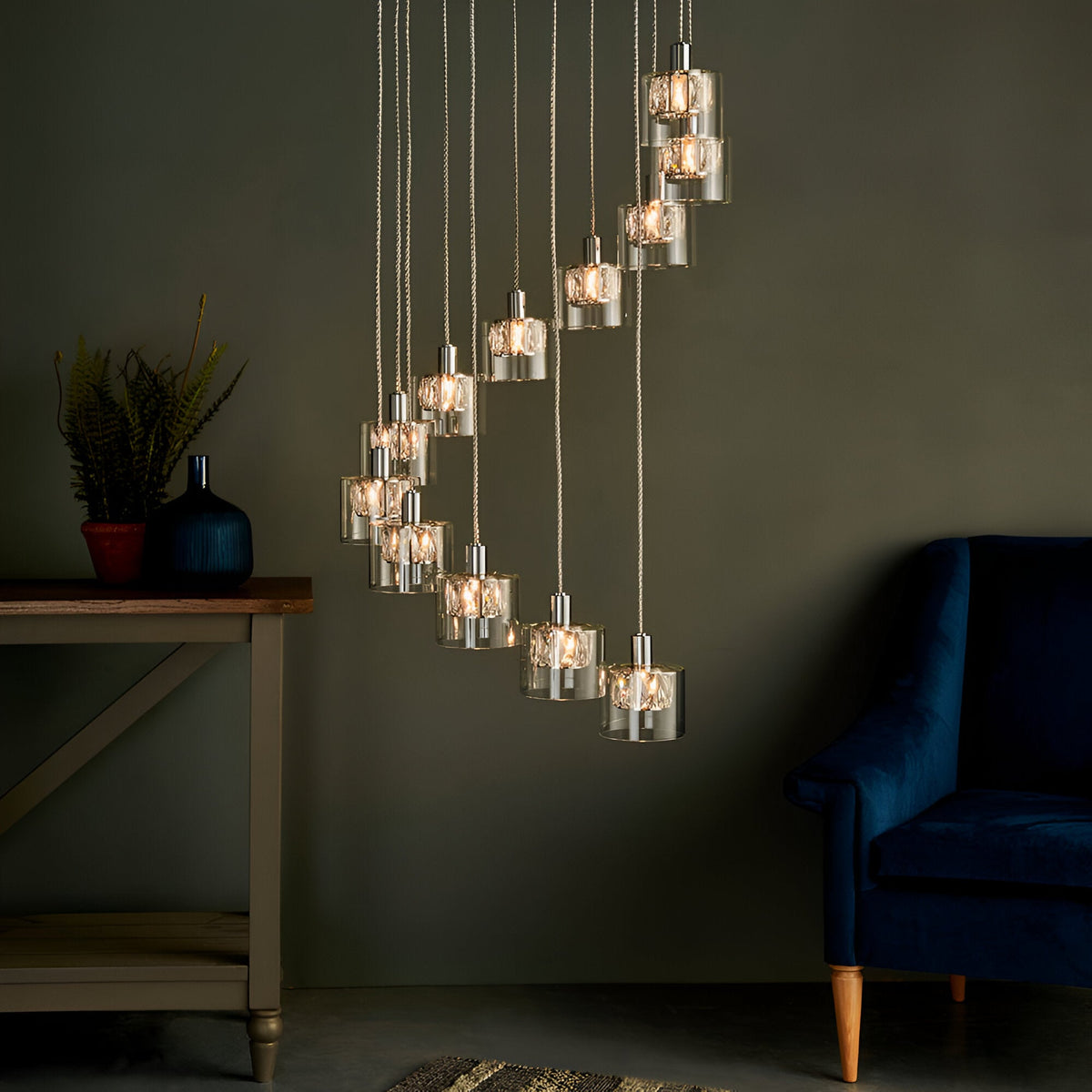 Marguerite 12-light pendant in polished chrome with cascading crystal-filled glass shades at staggered heights, lit above a console table in a dark living room.