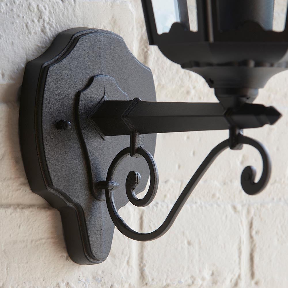 Close-up of Endon Ashshaw matt black outdoor wall lantern bracket, showing die-cast aluminium backplate and decorative scrollwork mounted on a white brick wall.