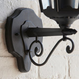 Close-up of Endon Ashshaw matt black outdoor wall lantern bracket, showing die-cast aluminium backplate and decorative scrollwork mounted on a white brick wall.