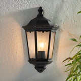 Ashshaw Coach Lantern in matt black die-cast aluminium with clear glass panels, mounted on a textured wall and lit with a visible bulb.