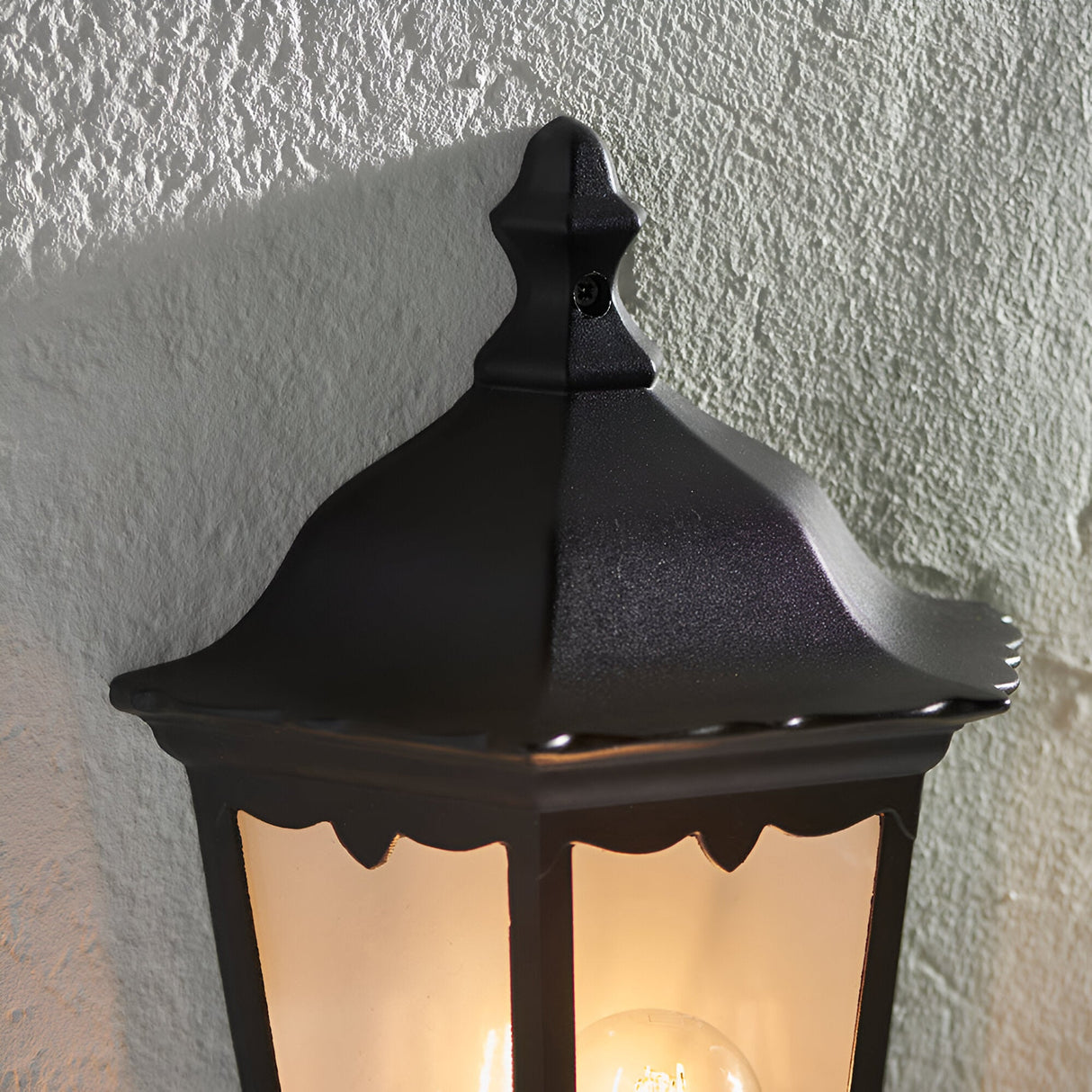 Ashshaw Coach Lantern in matt black die-cast aluminium, close-up of the peaked roof and clear glass panel with glowing bulb, mounted against a textured wall.