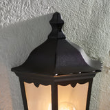 Ashshaw Coach Lantern in matt black die-cast aluminium, close-up of the peaked roof and clear glass panel with glowing bulb, mounted against a textured wall.