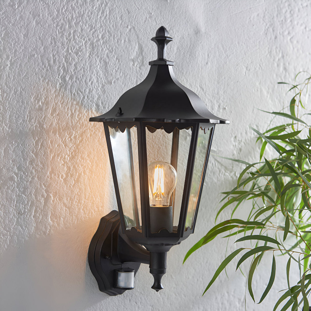 Endon Ashshaw traditional outdoor wall lantern in matt black die-cast aluminium with clear glass, E27 filament bulb and PIR sensor, on a white wall.