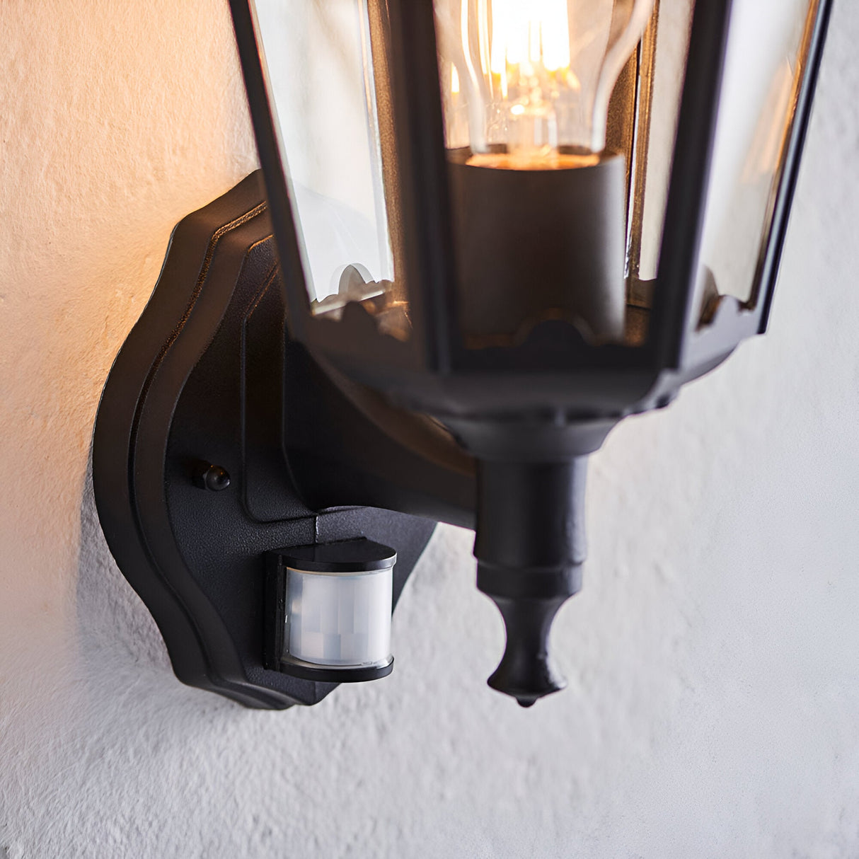 Endon Ashshaw outdoor wall lantern in matt black die-cast aluminium with clear glass panels, lit E27 filament bulb and PIR sensor, mounted on a pale wall.