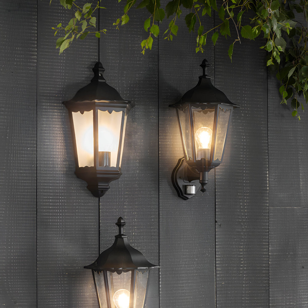Endon Ashshaw traditional outdoor wall lanterns in matt black aluminium with clear glass panels, one featuring a PIR sensor, mounted on grey wood cladding.