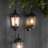 Endon Ashshaw traditional outdoor wall lanterns in matt black aluminium with clear glass panels, one featuring a PIR sensor, mounted on grey wood cladding.