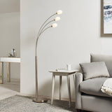 Endon Brockcombe 3 light floor lamp in satin nickel with white glass shades on arched arms, beside a side table and grey sofa in a living room.