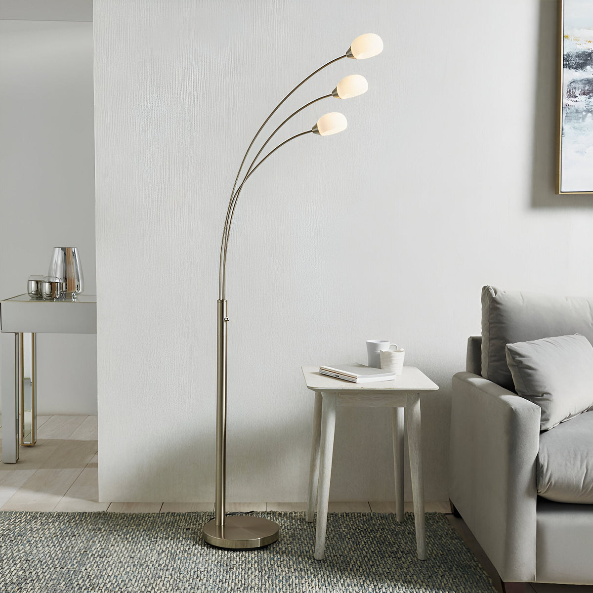 Endon Brockcombe 3 light floor lamp in satin nickel with white glass shades on arched arms, beside a sofa and side table in a modern living room.