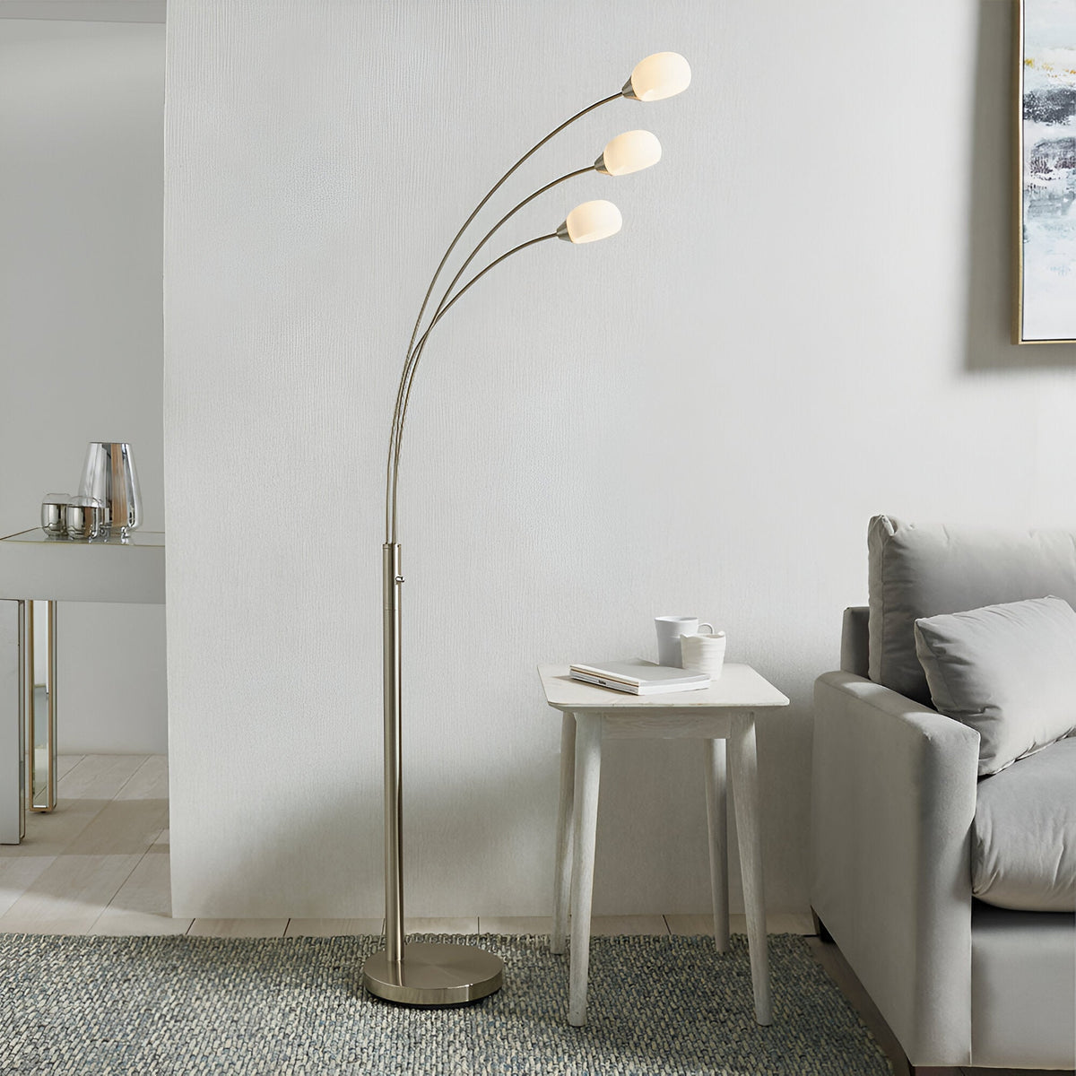 Brockcombe floor lamp with three arched satin nickel arms, each topped with a white glass shade, shown beside a sofa in a living room.