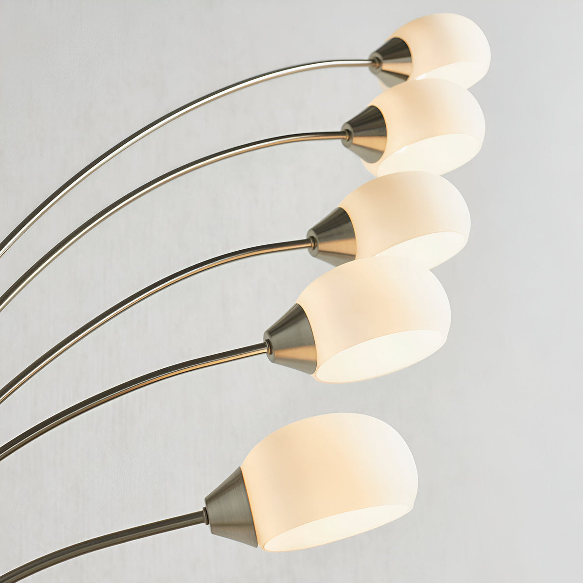 Brockcombe 5 Light Floor Lamp in satin nickel, close-up of arched arms with white glass shades, lit warm white against a light grey background.