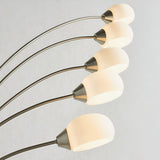 Brockcombe 5 Light Floor Lamp in satin nickel, close-up of arched arms with white glass shades, lit warm white against a light grey background.
