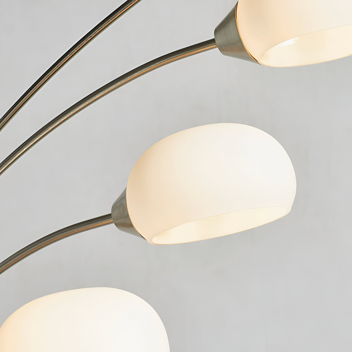 Close-up of the Brockcombe 5 Light Floor Lamp's satin nickel arched arms and white glass shades, one lit, against a plain grey background.