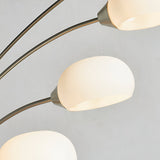 Close-up of the Brockcombe 5 Light Floor Lamp's satin nickel arched arms and white glass shades, one lit, against a plain grey background.