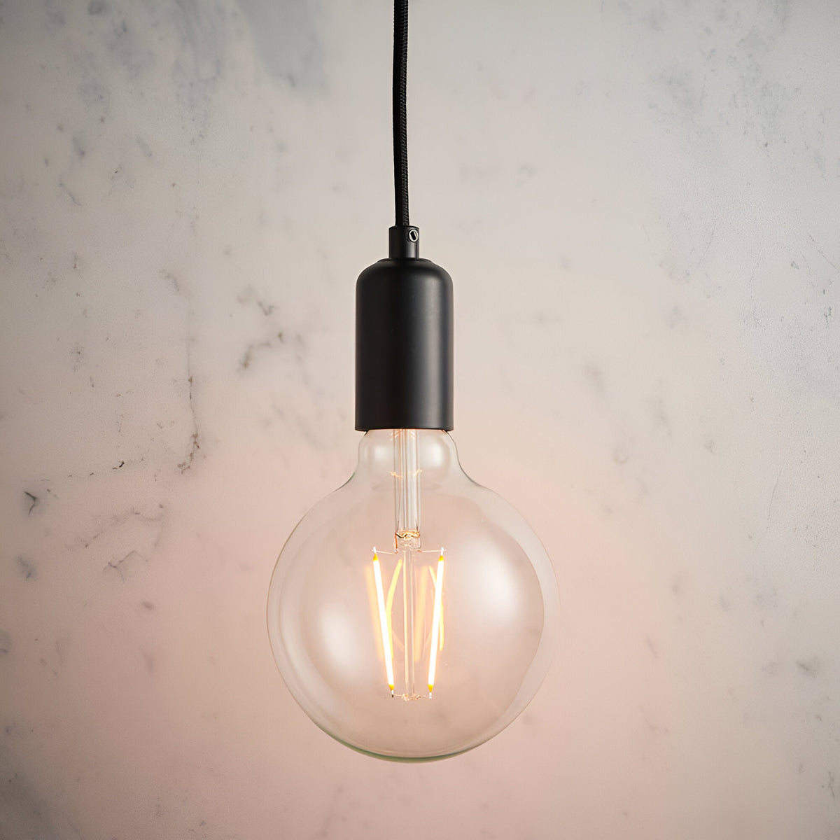 Endon Marlowe pendant light with matt black E27 lampholder on black fabric flex, fitted with an exposed globe filament LED bulb against a marble wall.