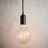 Endon Marlowe pendant light with matt black E27 lampholder on black fabric flex, fitted with an exposed globe filament LED bulb against a marble wall.