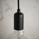 Close-up of the Endon Marlowe matt black E27 lampholder on black fabric flex, with a clear filament bulb below against a marble backdrop.