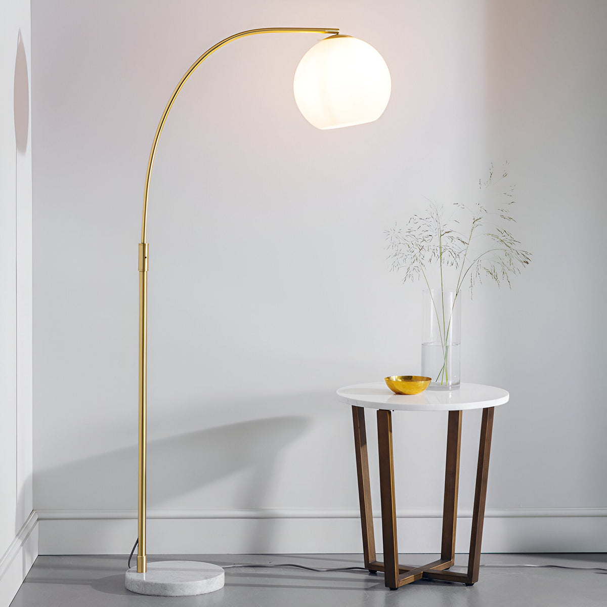 Endon Sinclair floor lamp with arched brass stem, gloss white glass globe shade and marble base, beside a round side table in a light interior.