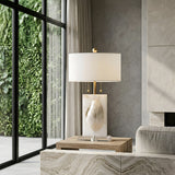 The Carved 1 Light Alabaster Table Lamp – Brass & Soft White is displayed on a wooden side table beside a beige sofa, with large windows and a lush vertical garden enhancing the bright, modern living space.