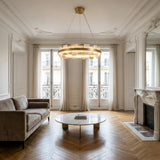 Elegant living room with a beige sofa, round marble coffee table, herringbone wood floor, ornate fireplace, tall windows with sheer curtains, and the Montyra LED Large Panelled Alabaster Chandelier in soft white & brass.