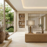A modern, minimalist living room with large windows, neutral tones, a wooden table topped with the Elvaris 1 Light Framed Marble Table Lamp in brass & grey and books, built-in shelves with decor, potted plants, and a view of a small green garden outside.