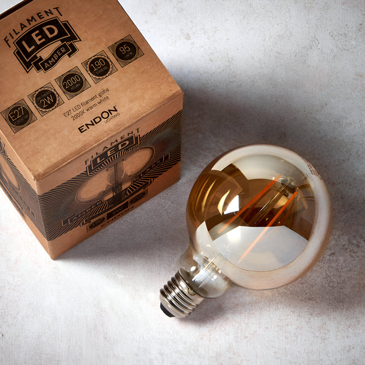 2W E27 LED filament globe bulb with amber tinted glass, visible orange filament, warm white 2200K, next to its Filament LED Amber packaging.