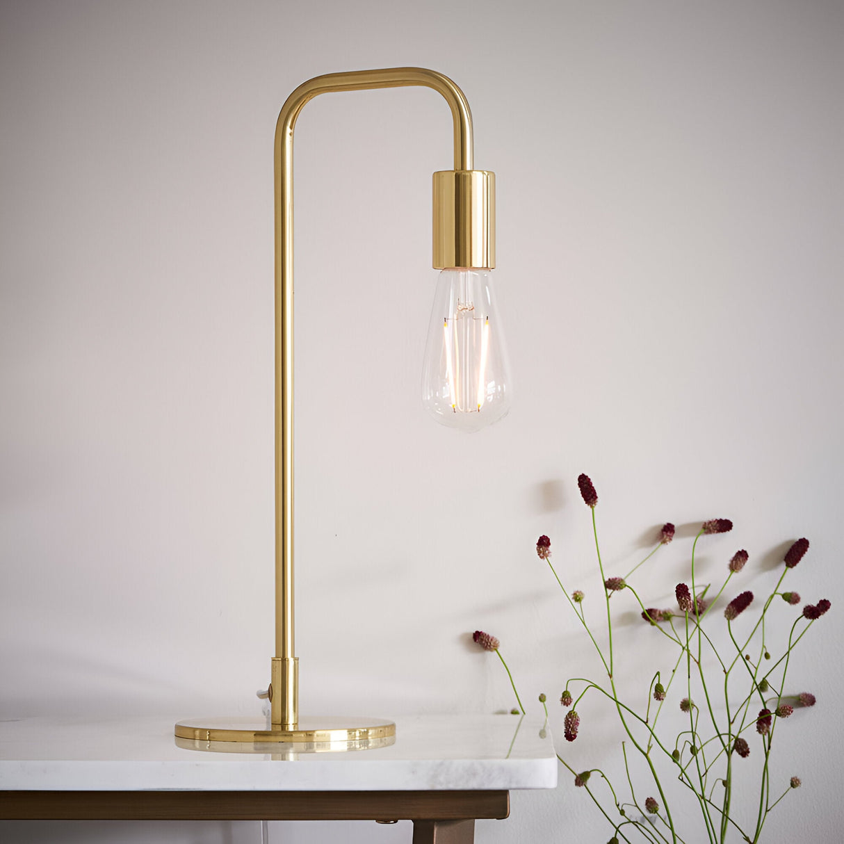 Endon Cliffgrove brushed brass table lamp with slim arched frame and exposed filament bulb, shown on a marble surface beside dried flowers.