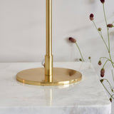 Close-up of the Endon Cliffgrove table lamp's round brushed brass base and slim stem on a marble surface beside dried pink flowers.