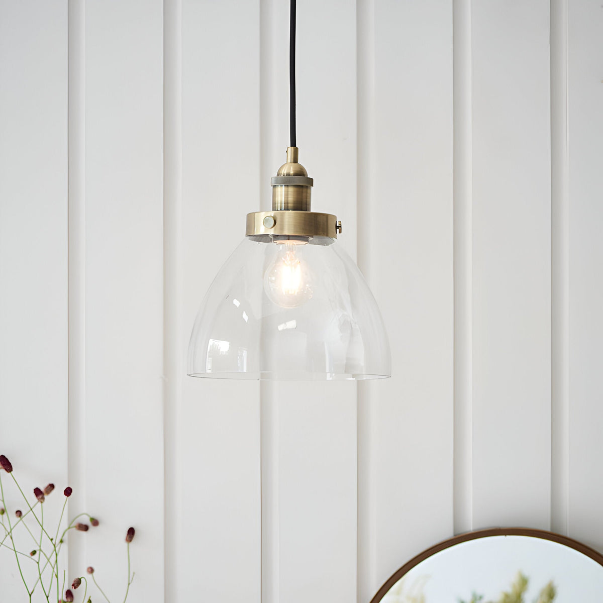 Endon Wentworth industrial pendant light with clear glass dome shade, knurled brass lamp holder and black flex, hung against white panelled wall.