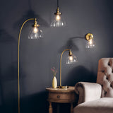 Endon Wentworth pendant light in brass with clear glass shade shown alongside matching floor lamp, wall light and table lamp in a dark living room setting.