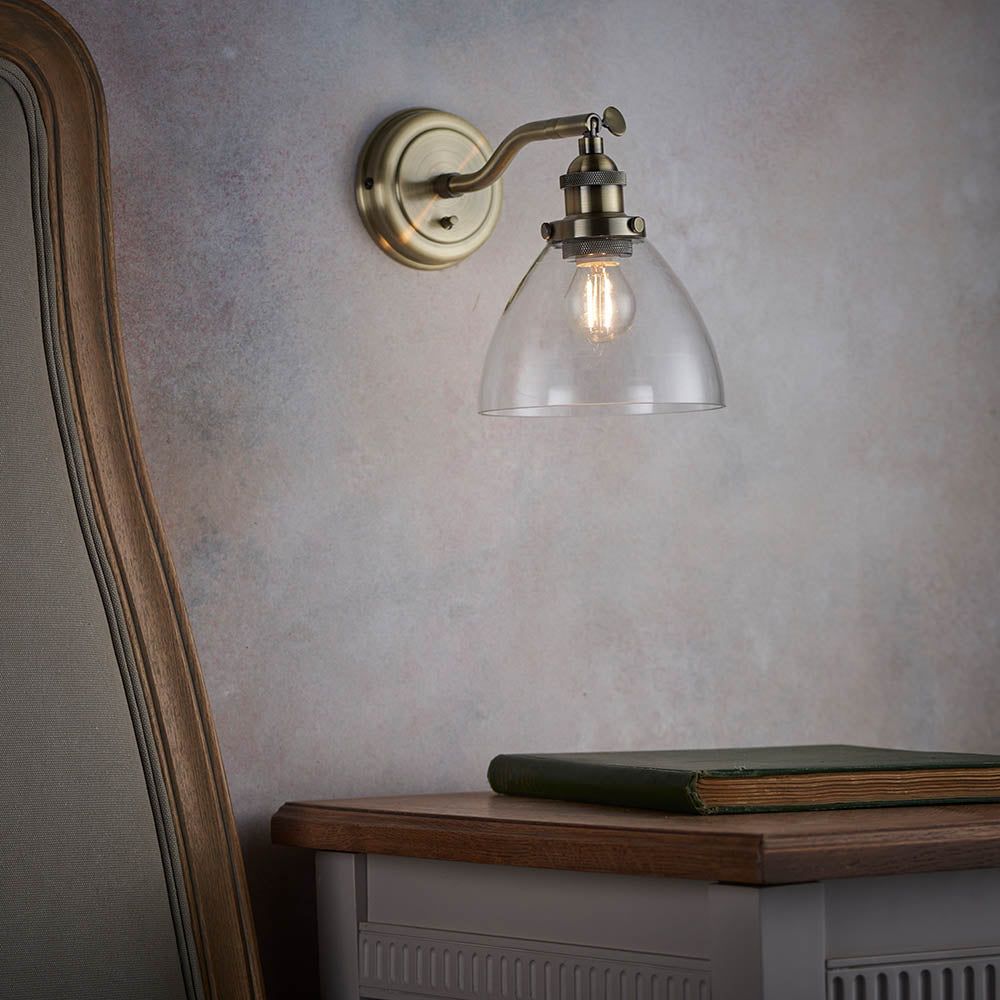 Wentworth Wall Light in brass with an adjustable knuckle arm and clear glass dome shade, mounted above a wooden bedside table in a bedroom setting.