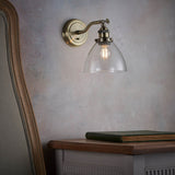 Wentworth Wall Light in brass with an adjustable knuckle arm and clear glass dome shade, mounted above a wooden bedside table in a bedroom setting.