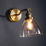 Wentworth Wall Light in brass with adjustable knuckle joint, knurled lamp holder and clear glass dome shade, shown lit against a dark grey wall.