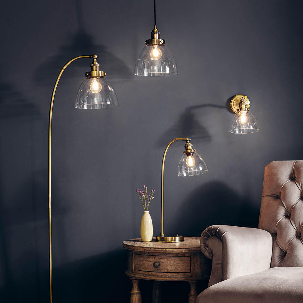 Wentworth Wall Light in brass with clear glass dome shade and adjustable knuckle joint, shown mounted on a dark grey wall in a living room setting.