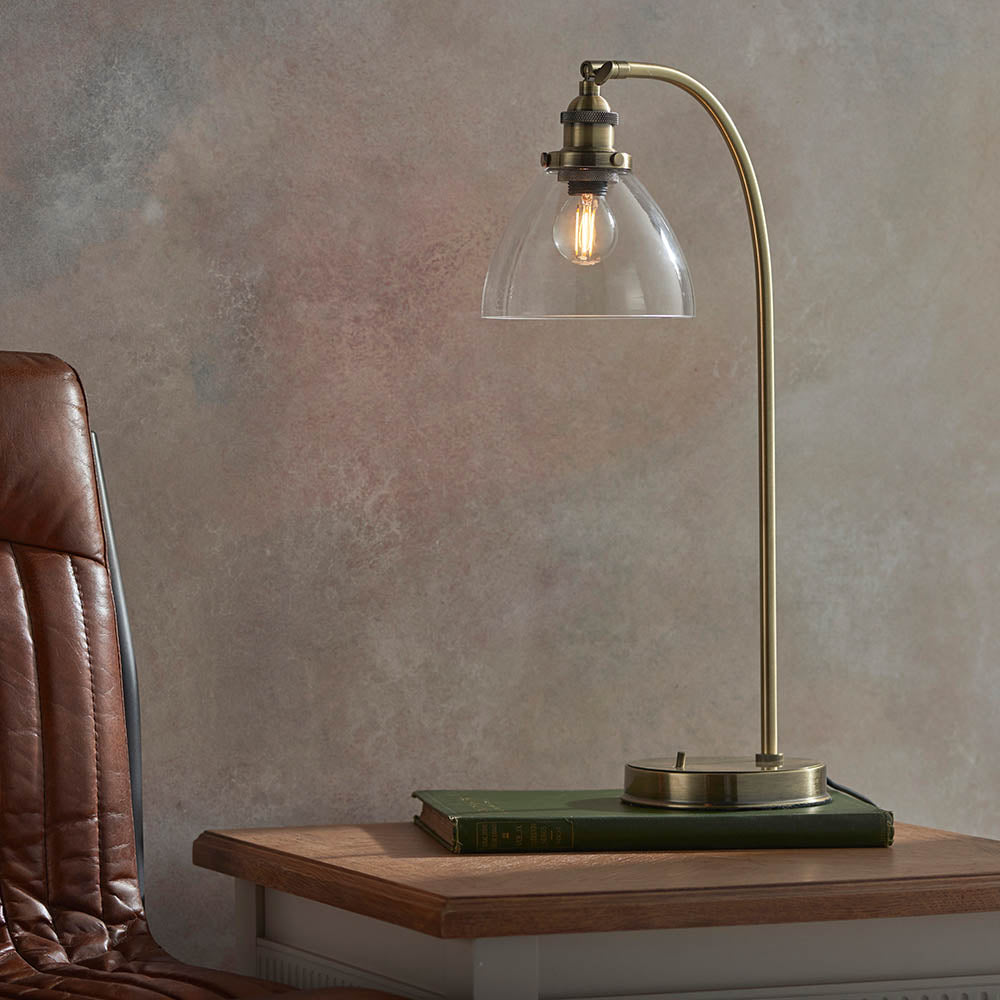 Wentworth Table Lamp in brass with clear glass dome shade and curved arm, shown on a wooden side table beside a leather chair.