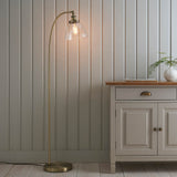 Endon Wentworth arched floor lamp in antique brass with a clear glass dome shade and exposed filament bulb, beside a pale sideboard and panelled wall.