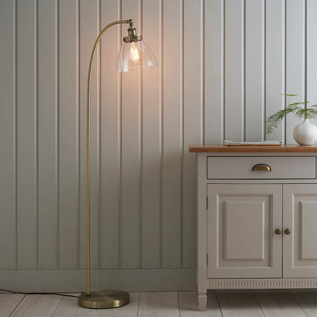 Endon Wentworth arched floor lamp in antique brass with a clear glass dome shade and exposed filament bulb, beside a pale sideboard and panelled wall.