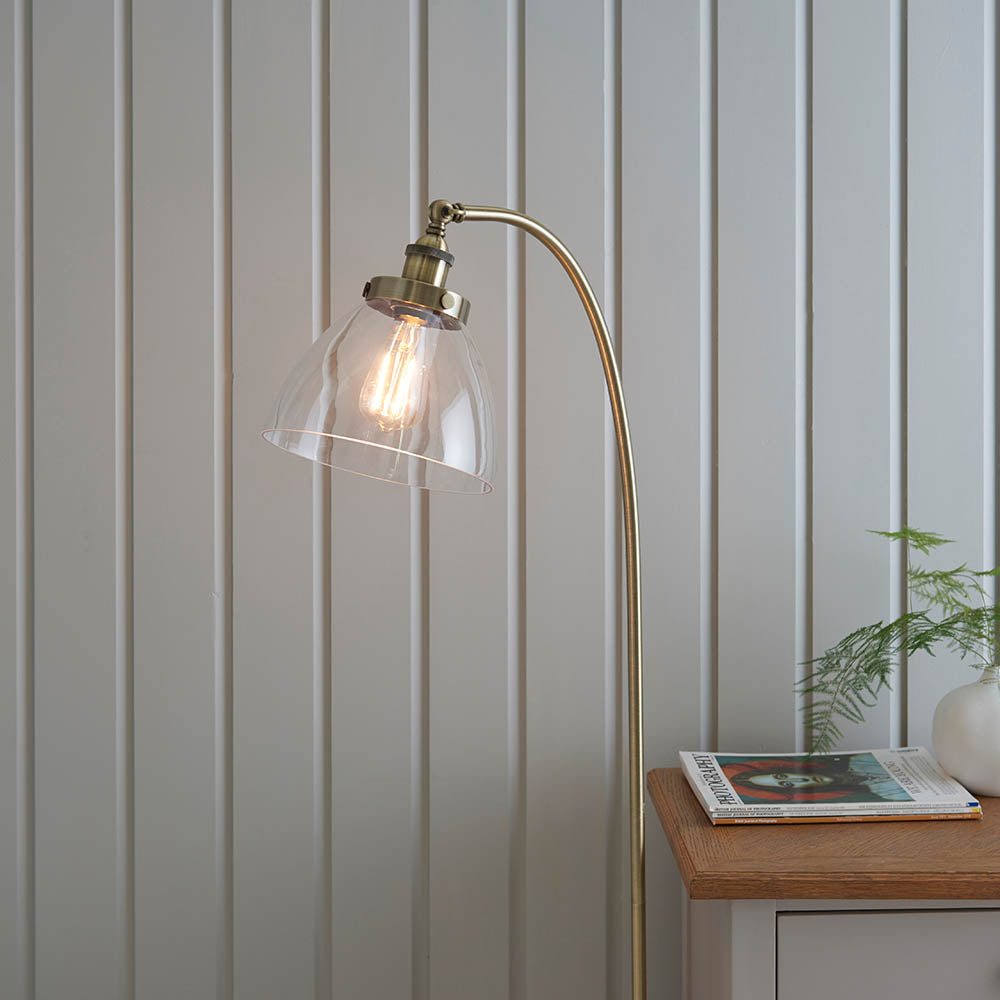 Endon Wentworth brass floor lamp with arched stem, clear glass shade and exposed filament bulb, beside an oak side table against pale panelled wall.