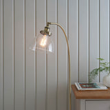 Endon Wentworth brass floor lamp with arched stem, clear glass shade and exposed filament bulb, beside an oak side table against pale panelled wall.