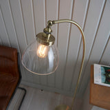 Endon Wentworth floor lamp in brass with adjustable knuckle, knurled lamp holder and clear glass shade lit by a filament bulb, beside a leather chair.