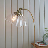 Endon Wentworth floor lamp in brass with an arched stem and tilted clear glass shade holding a visible filament bulb, beside a light panelled wall.